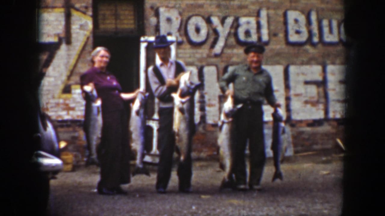 In 1939 Wyoming home-movie footage, a woman and two men pose with large fish before a brick wall bearing a faded Royal Blue sign.