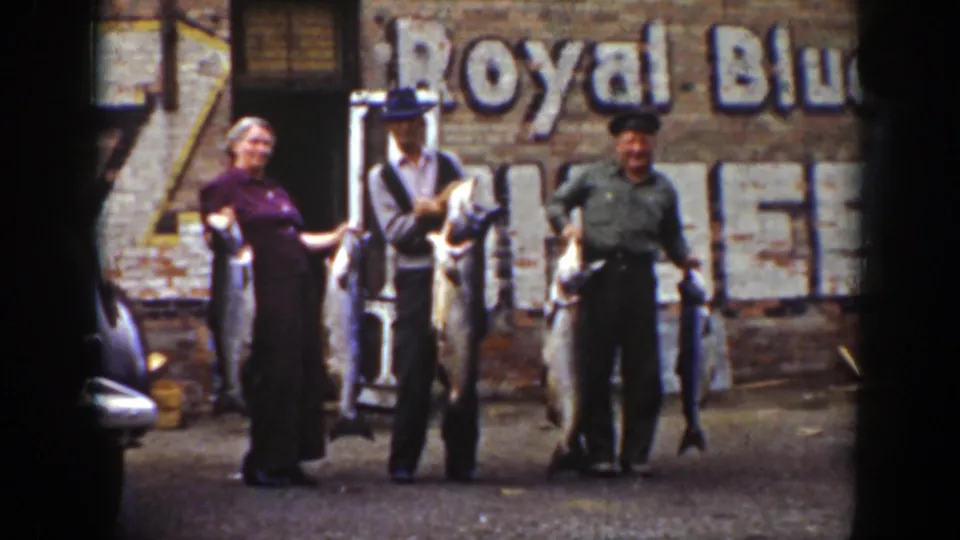 In 1939 Wyoming home-movie footage, a woman and two men pose with large fish before a brick wall bearing a faded Royal Blue sign.