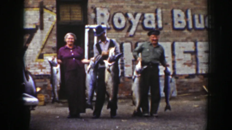 A woman and two men smile while holding their catch, framed by a brick wall with faded Royal Blue advertising text.