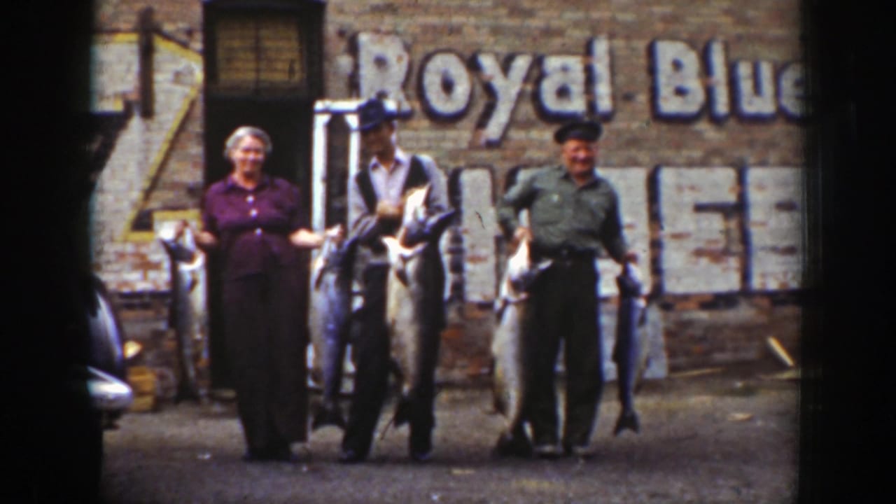 In Wyoming 1939 home-movie footage, adults pose with large fish in front of a brick wall with faded Royal Blue lettering.
