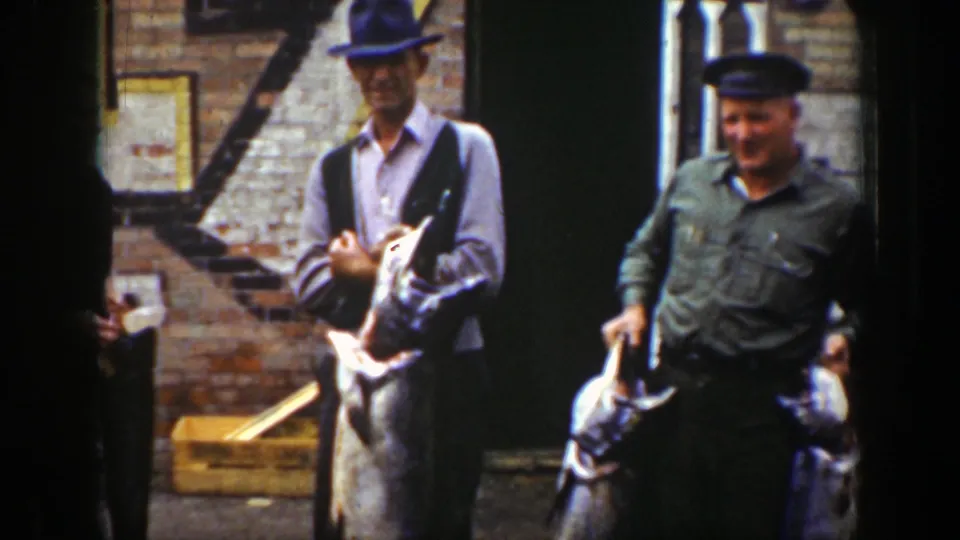 Two men, one in a cap and one in a green shirt, hold large fish near a brick storefront doorway in 1939 Wyoming footage.