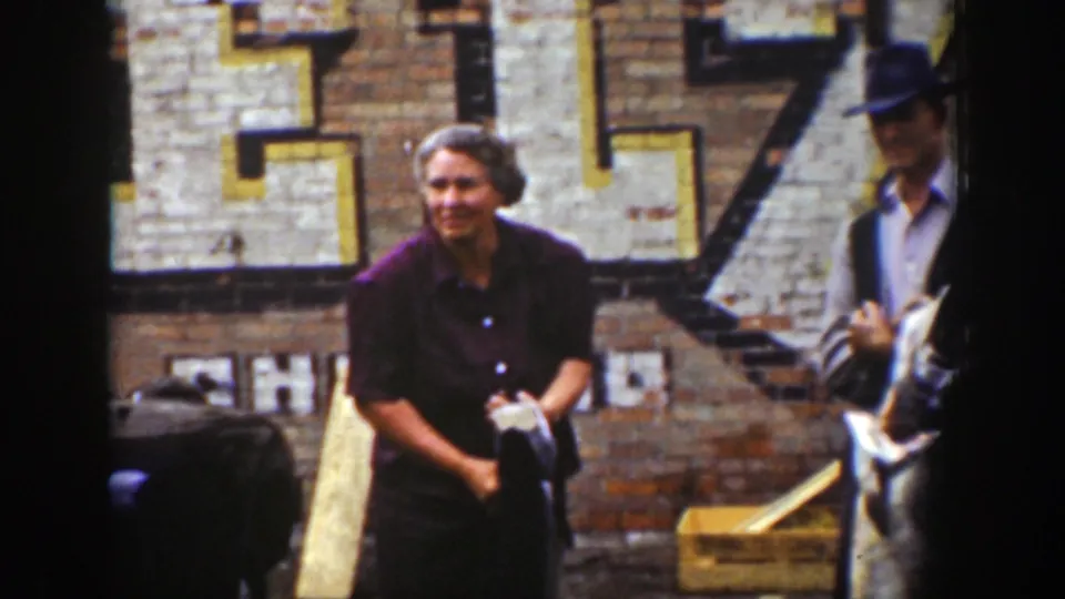 A woman in a purple dress smiles with a large fish in front of faded storefront lettering in Wyoming 1939 home-movie footage.
