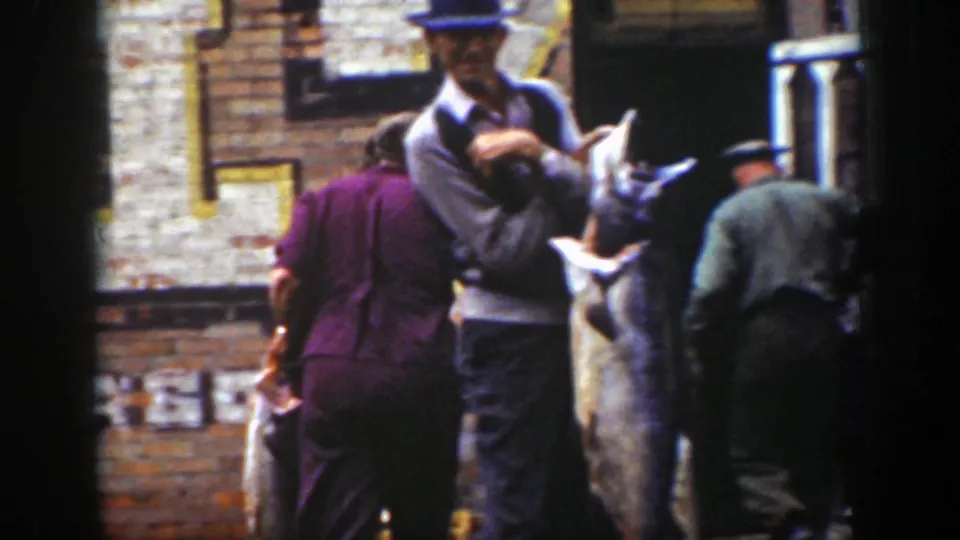 A man in a hat displays a large fish outside a brick building, with other adults gathered close in the Wyoming scene.
