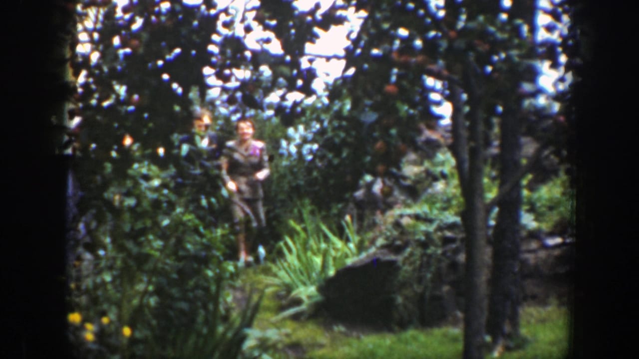 Two adults stand among dense garden greenery and trees.