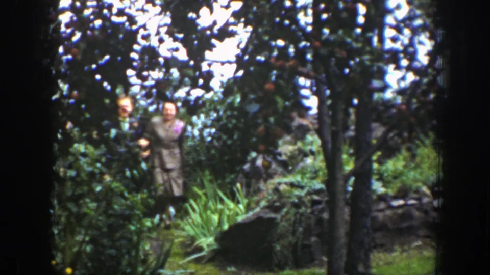 A man and woman walk through a green Wyoming garden in 1939, moving toward the camera through trees and plants.