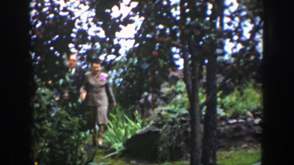 A woman in a dress steps through the Wyoming garden, with another adult partly visible behind her among the trees.