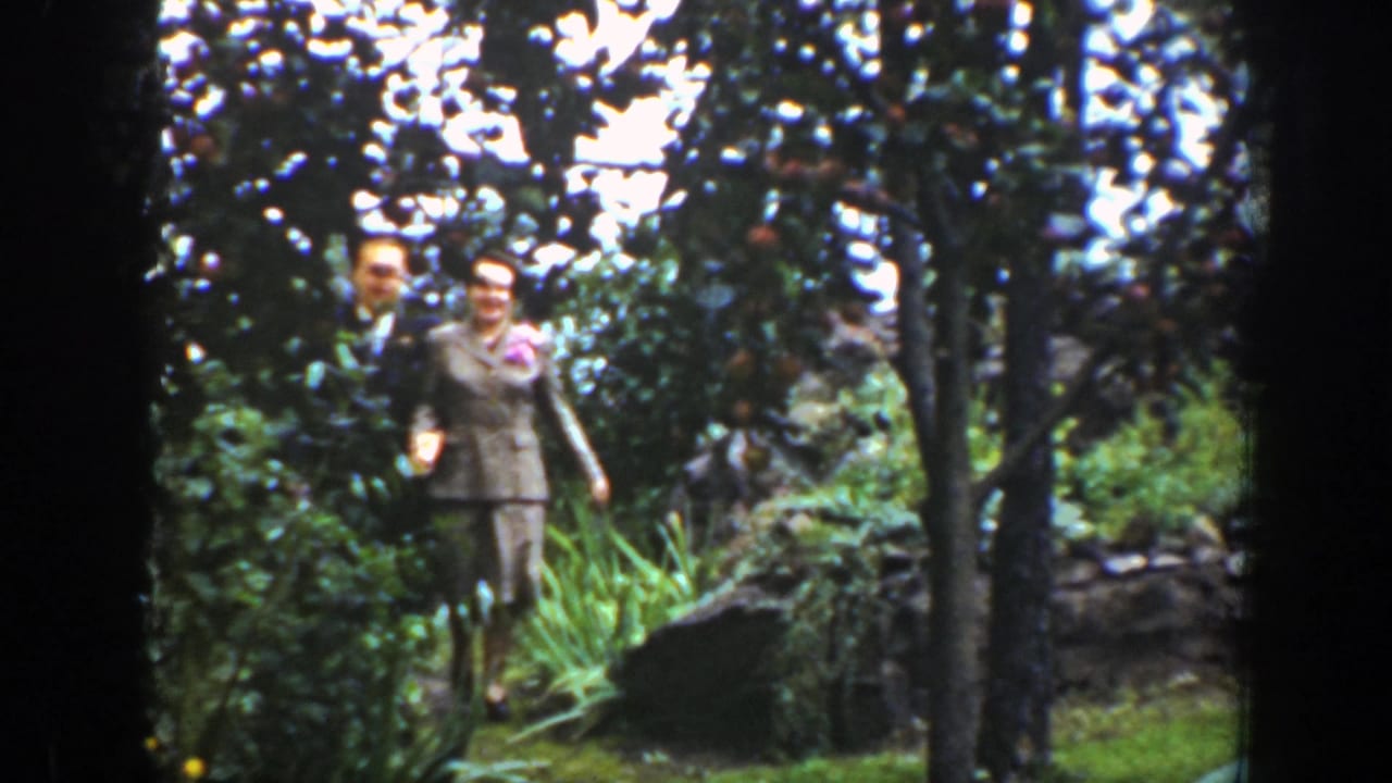Well-dressed couple walking arm in arm through a leafy garden.