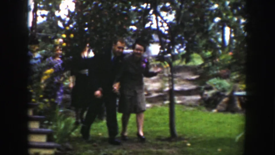 The suited pair walks across the garden lawn beneath fruit trees, with stonework and dense greenery behind them.
