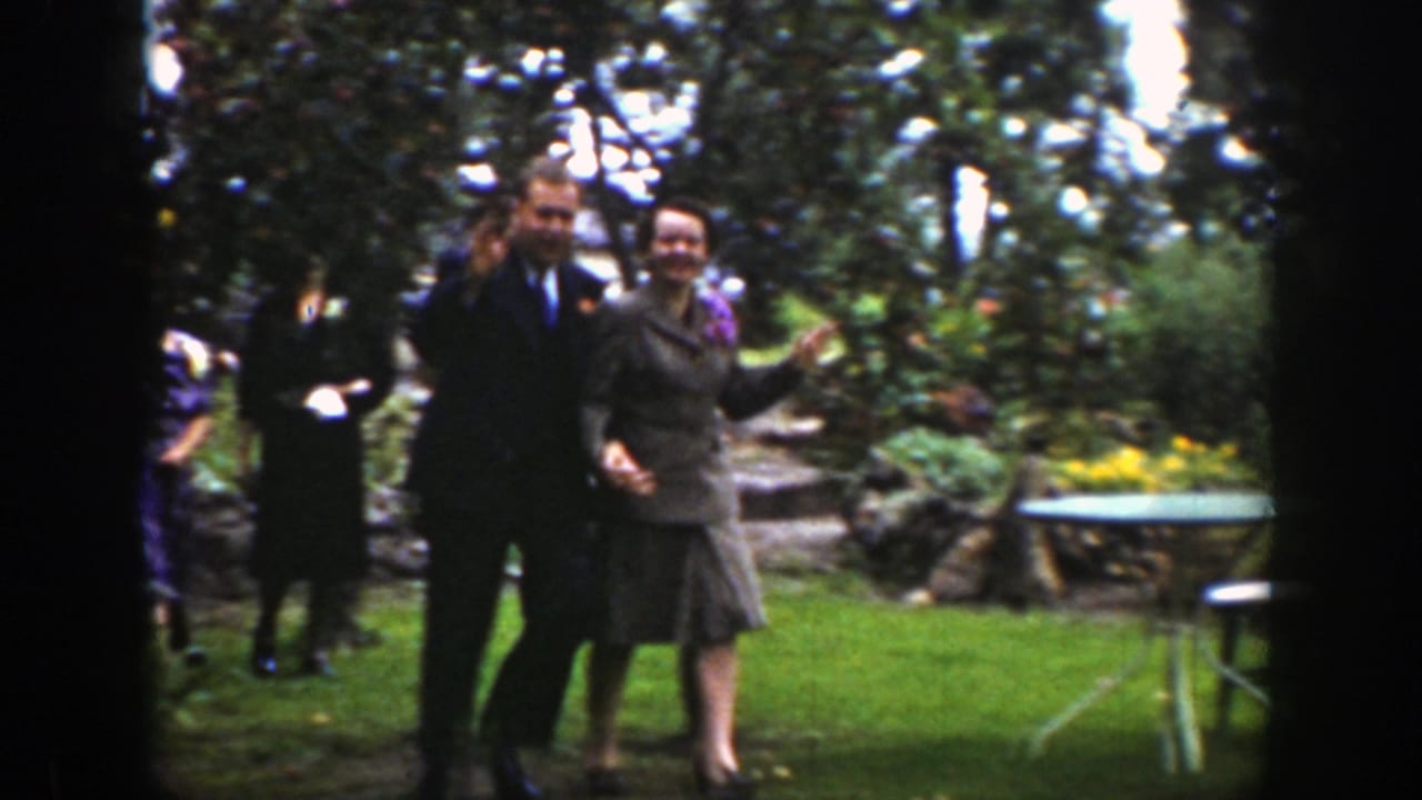 Adults in formal clothing walk through a garden, with a couple holding hands near the camera.