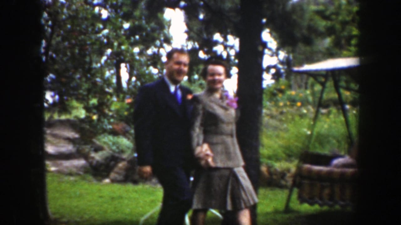 A man and woman hold hands and smile while walking past trees and garden furniture.