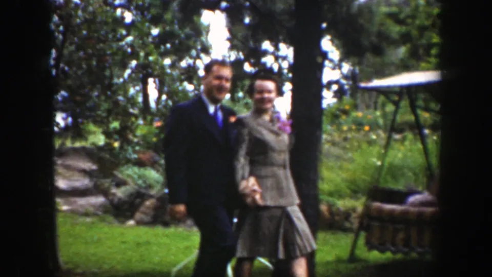 The couple smiles toward the camera as they walk hand in hand through a Wyoming garden, with a small table and trees behind them.