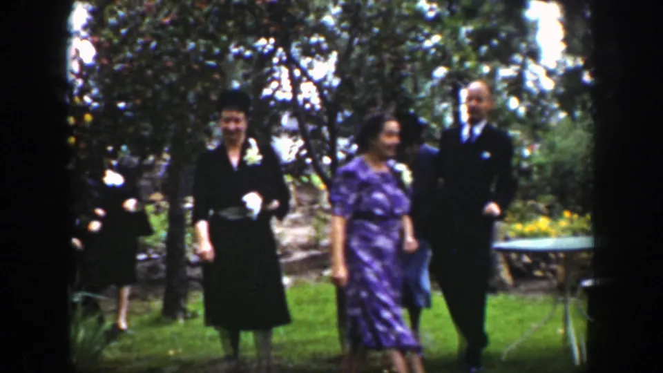 Adult guests in period dress walk across the garden in Wyoming, with trees, flowers, lawn, and a small table nearby.