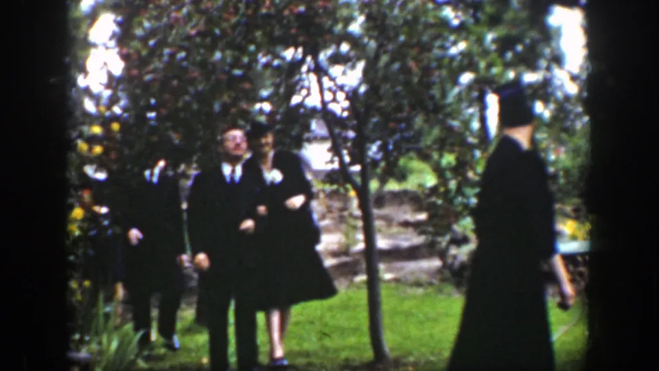 In the Wyoming garden-party scene, a formally dressed pair walks together as another guest passes in the foreground.