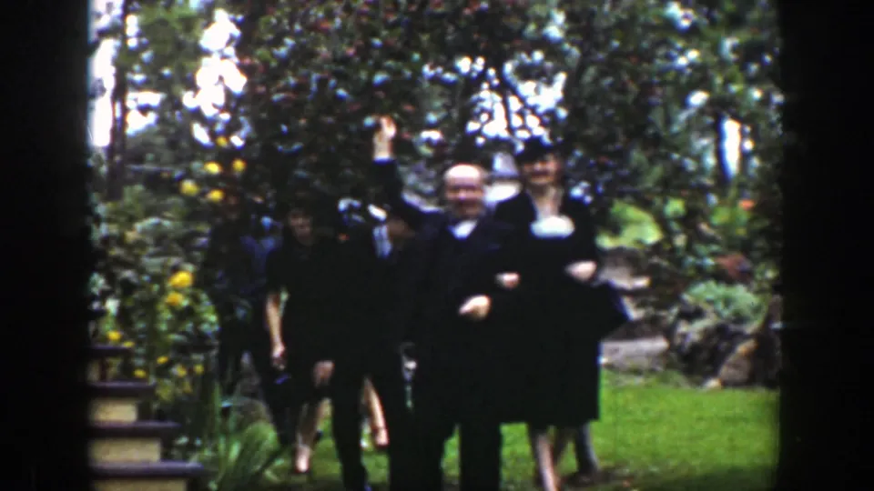 A male guest waves toward the home-movie camera as other formally dressed adults approach through the 1939 Wyoming garden setting.