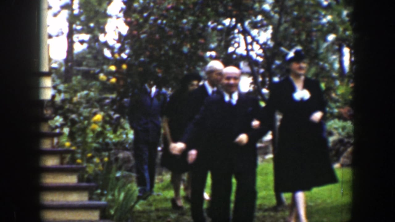 Adults in formal clothing stand and walk through a garden with trees and a house edge nearby.