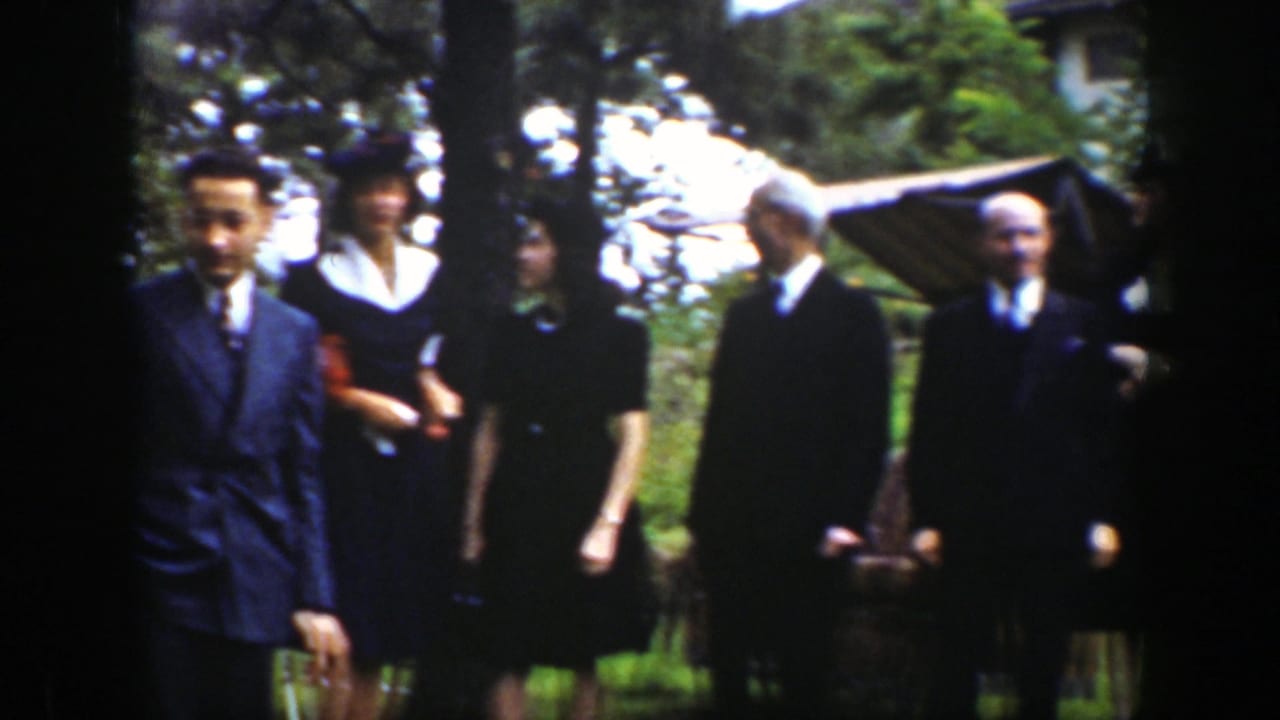 Several adults in suits and dark dresses stand near trees and a striped canopy.