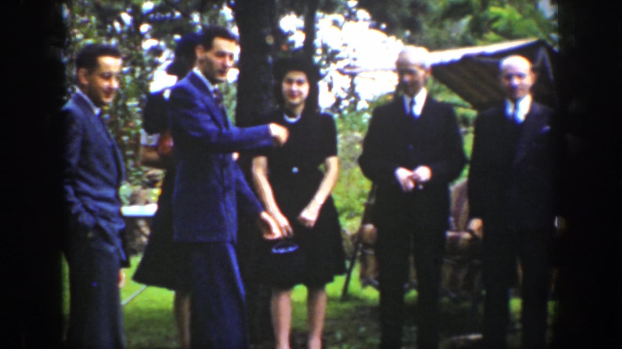 A suited man gestures while adults stand in a garden setting.
