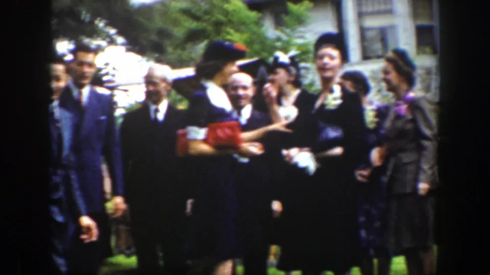 A woman in a dark hat appears to greet or embrace someone among formally dressed adults outdoors.