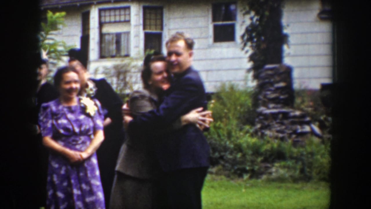 Adults stand on a lawn as a man in a dark suit hugs a woman near a white house.