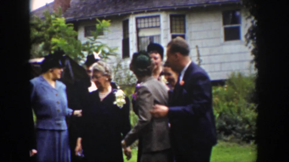 1939 Wyoming frame of a suited man and several adults in dresses and hats gathered on a lawn in front of a clapboard house.