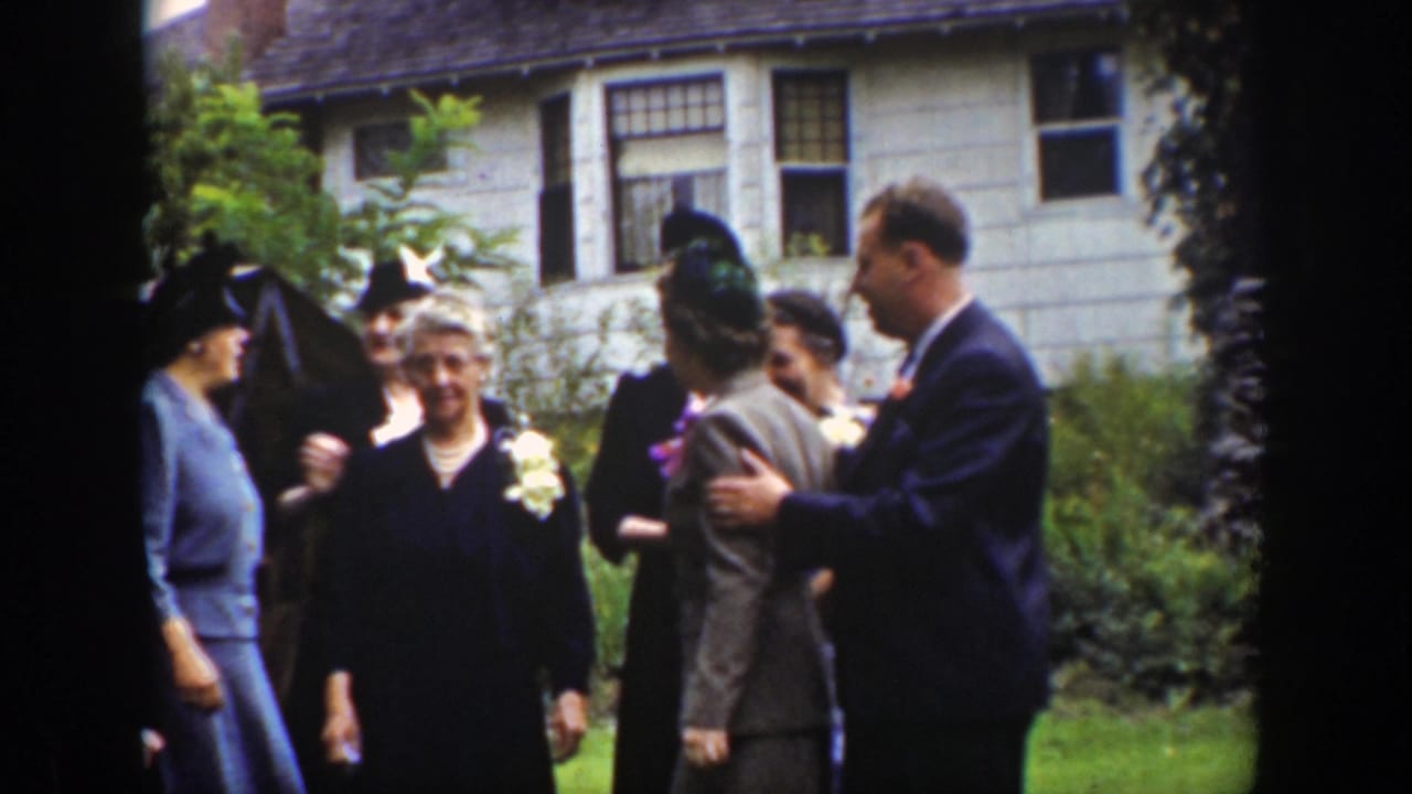 Adults in formal dress gather on a lawn outside a white house.