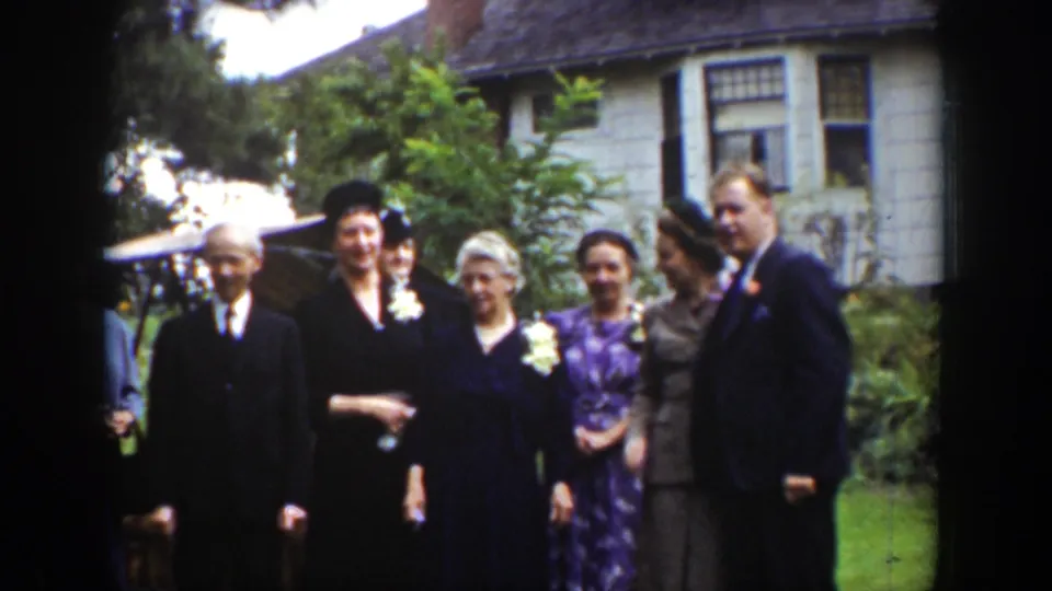 A 1939 Wyoming home-movie frame shows adults in formal clothing clustered on a lawn outside a house during a wedding-like gathering.