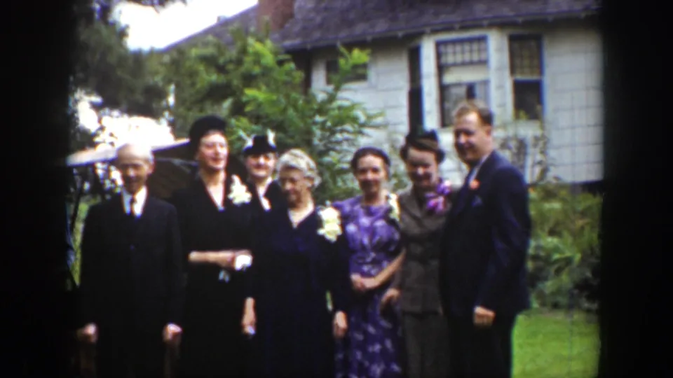 In Wyoming 1939 home-movie footage, adults in suits, hats, dresses, and corsages pose outdoors before a house.