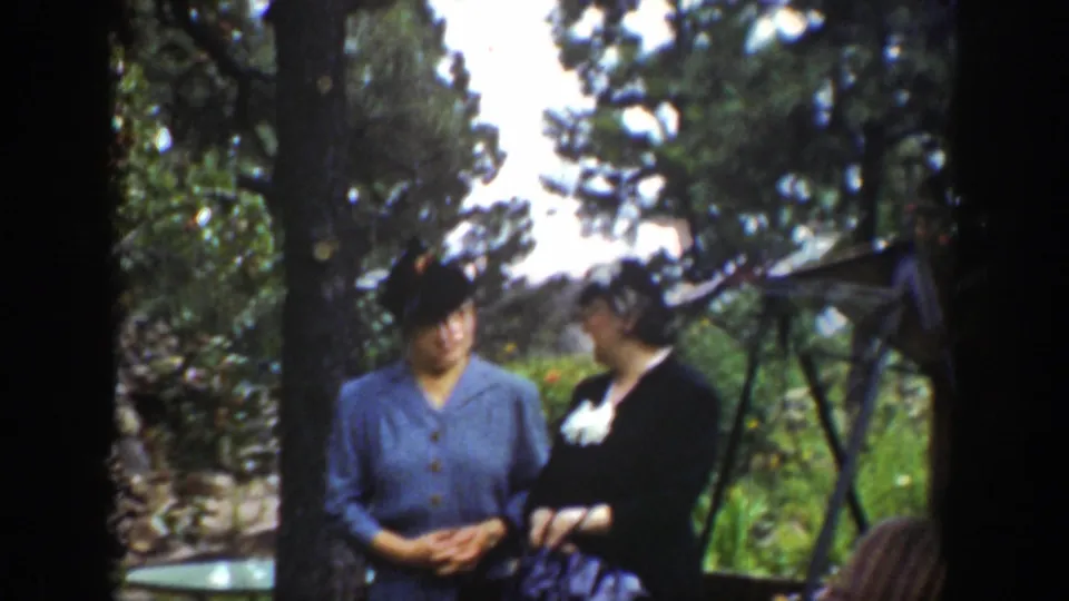 Two women in hats and formal dress converse outdoors in the Wyoming 1939 home-movie scene, framed by trees and garden greenery.