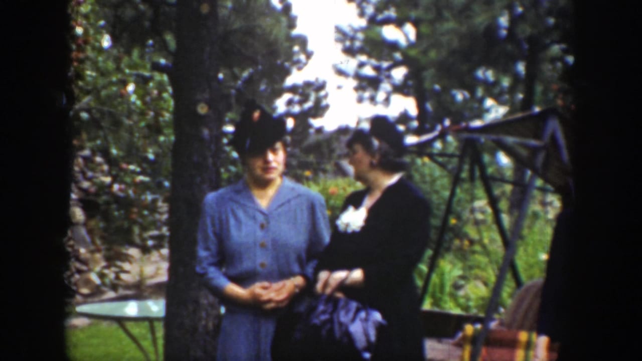 Two women in hats stand in a garden near trees and a lawn swing.