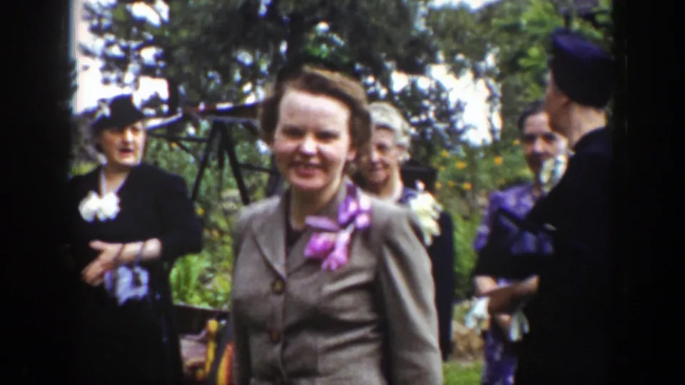 A group of adult women in period suits and hats gather outdoors, with one woman smiling toward the camera in the foreground.