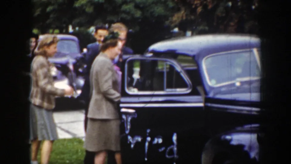 Adult women in period clothing gather beside a dark rounded sedan outdoors, with one woman near the open door area.