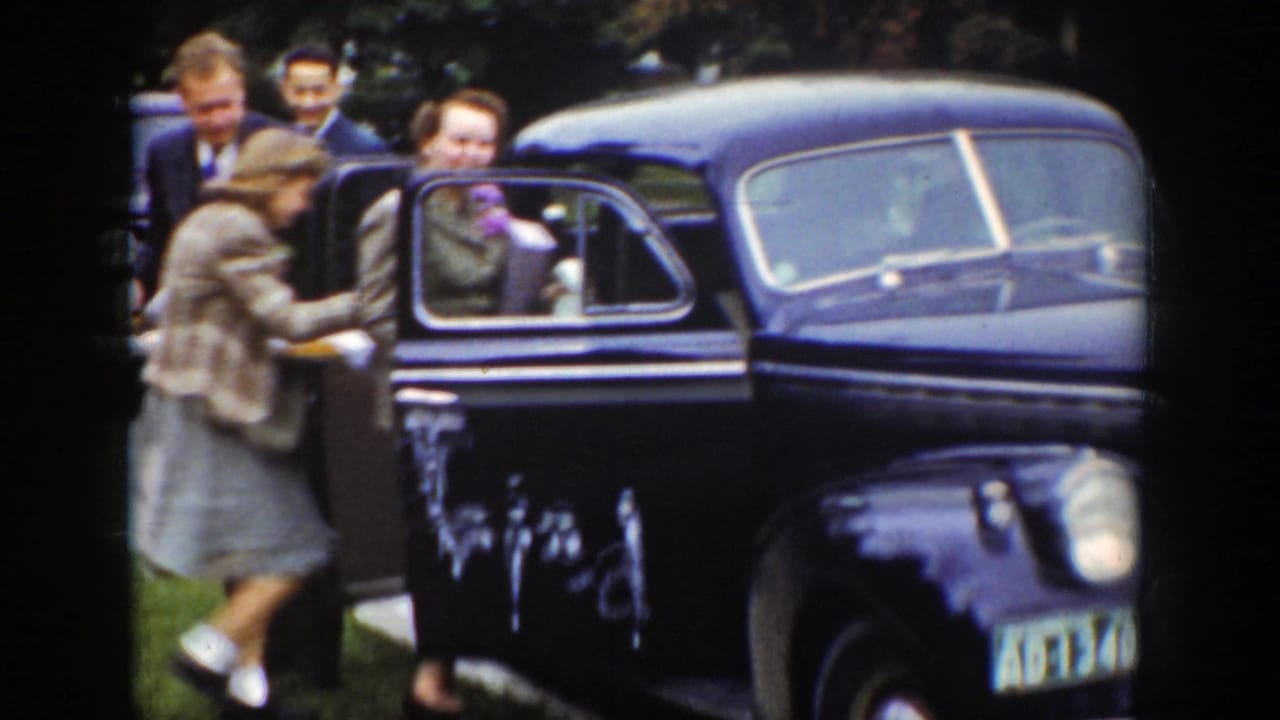 In 1939 Wyoming footage, a woman in a light coat and suited men gather beside an open door of a black sedan.