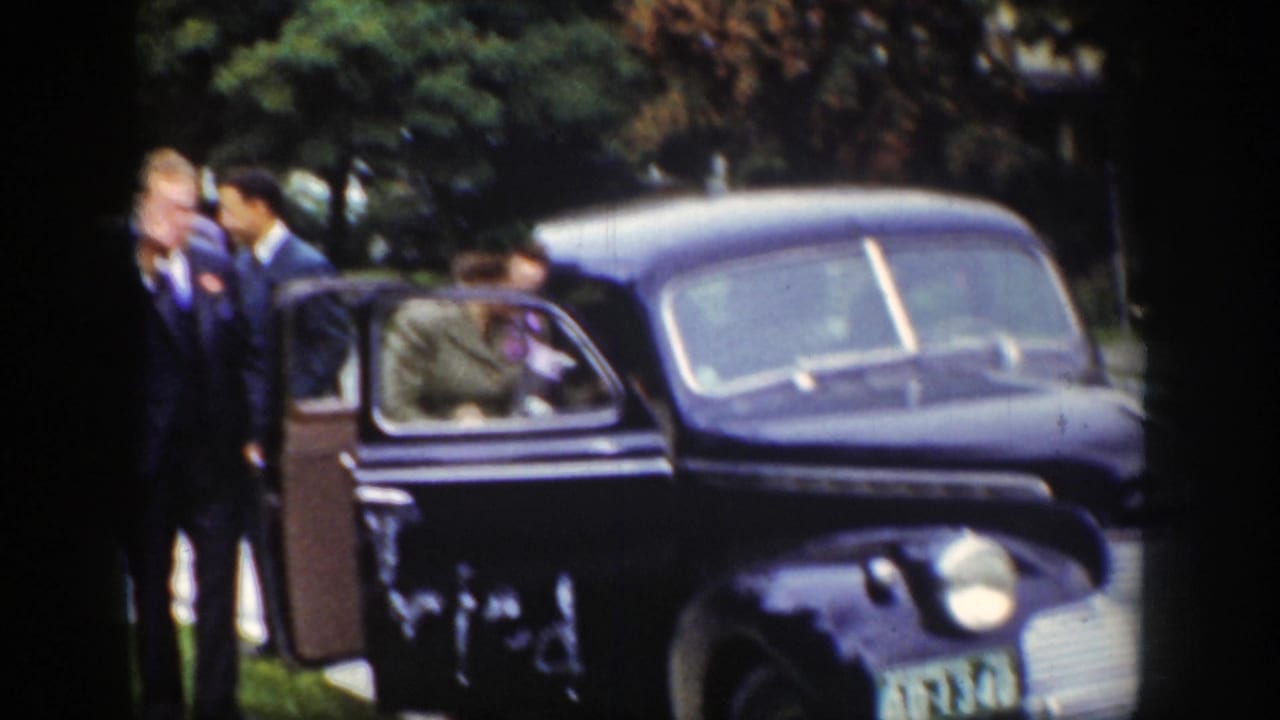 A group stands close to an open black sedan door with trees behind them.