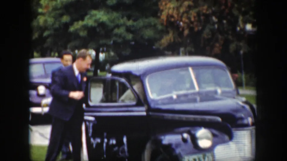 A man in a dark suit stands beside the open door of a black sedan, with another car visible behind him.