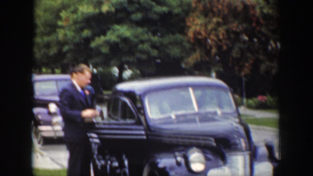 Man in a dark suit stands beside a decorated dark sedan on a residential street.