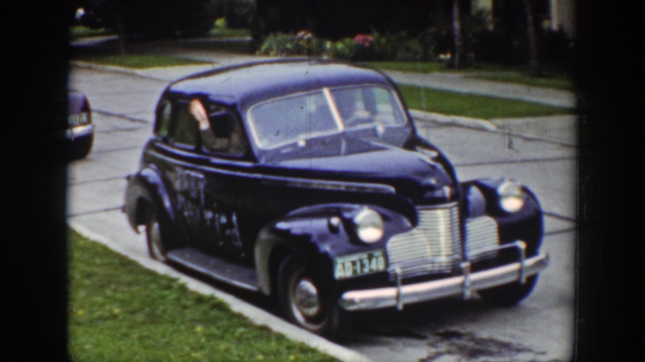 A dark blue 1939 Wyoming wedding sedan rolls along the curb while a passenger waves from the window.