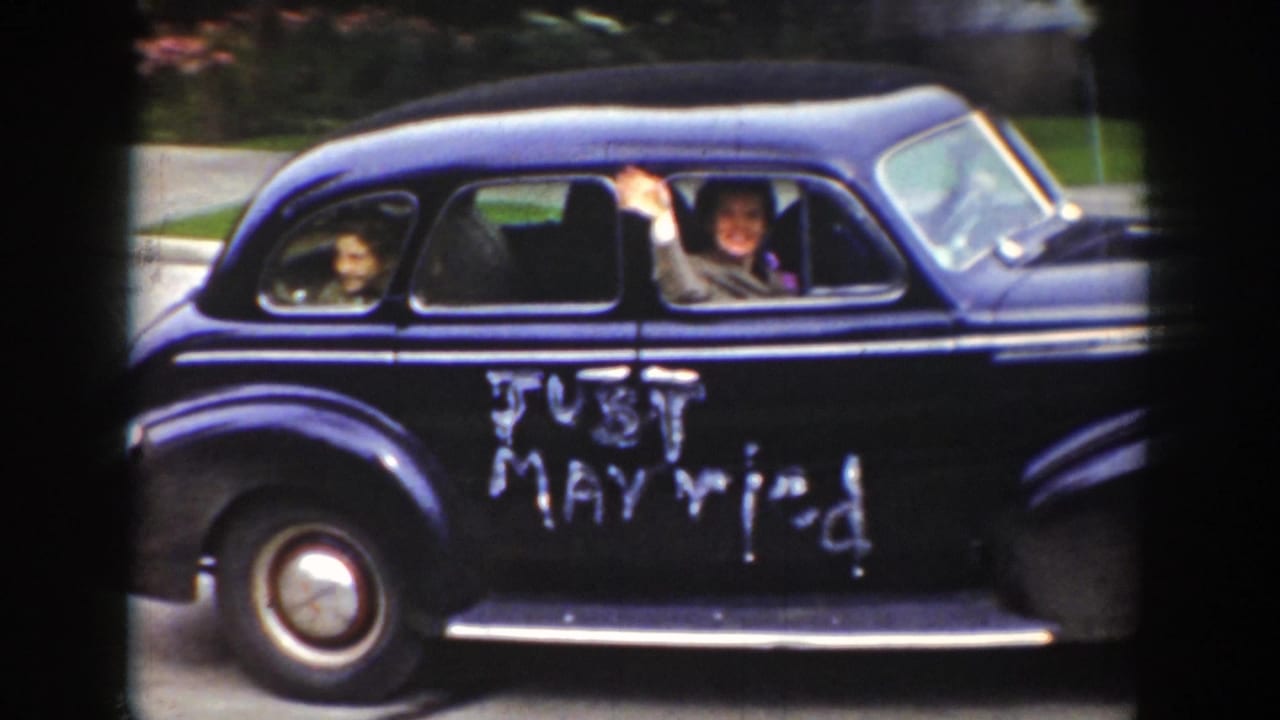 1939 Wyoming home-movie frame showing a dark wedding sedan moving past the camera with people visible through the windows and Just Married painted on the side.