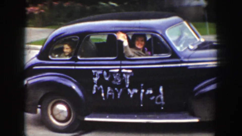 1939 Wyoming home-movie frame showing a dark wedding sedan moving past the camera with people visible through the windows and Just Married painted on the side.