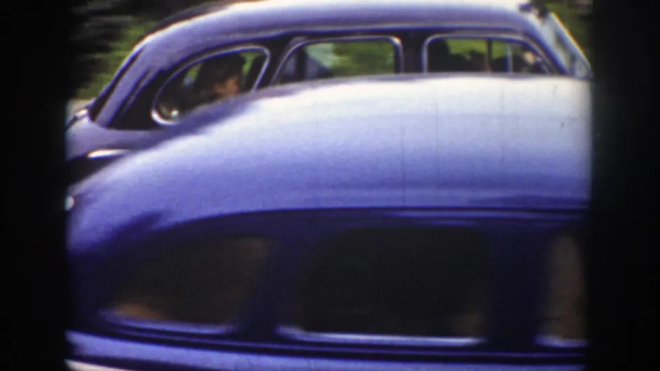 A soft 8mm frame shows the side of the wedding sedan with occupants faintly visible, partly blocked by a light-colored car crossing the foreground.