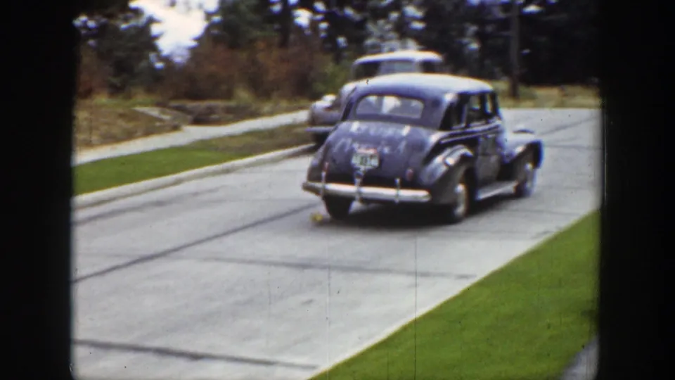 Rear view of the 1939 Wyoming wedding sedan driving away through a residential street, with another car ahead and lawns and trees along the road.
