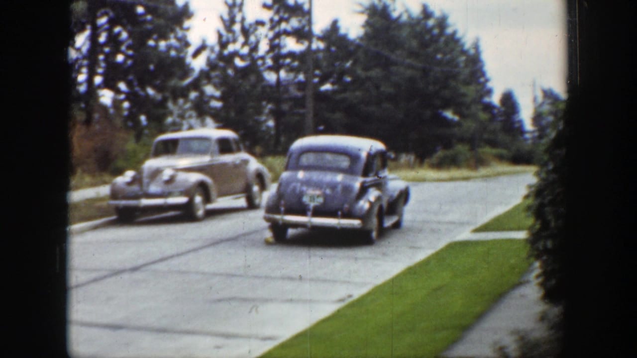1939 Wyoming home-movie frame with a light sedan and a darker car moving along a quiet paved street lined with grass and evergreens.