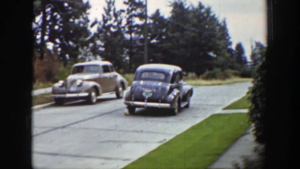 1939 Wyoming home-movie frame with a light sedan and a darker car moving along a quiet paved street lined with grass and evergreens.