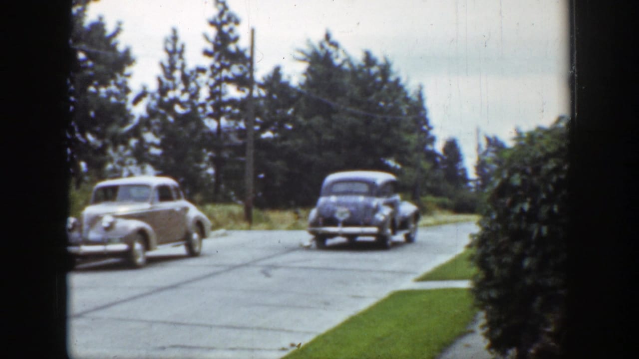 1939 Wyoming street scene showing a light sedan near the corner and a darker car moving away beside grass and tall evergreens.