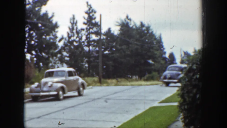 1939 Wyoming home-movie frame with a light sedan turning at the edge of the frame and a darker car receding along the quiet road.