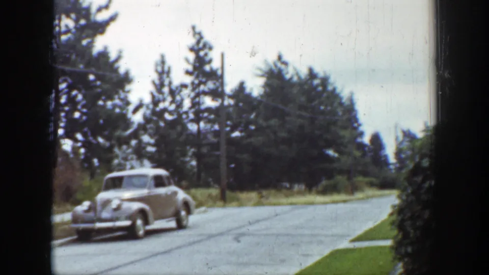 1939 Wyoming home-movie frame showing a light-colored sedan crossing the street foreground against grass, evergreens, and cloudy sky.