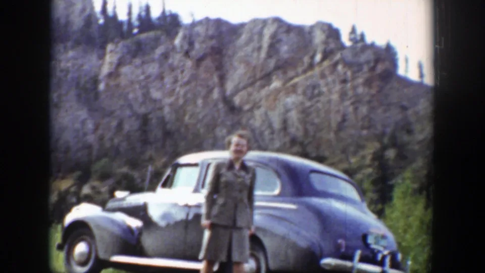 Woman in period clothing stands near a parked car beneath Wyoming cliffs in 1939 8mm footage.