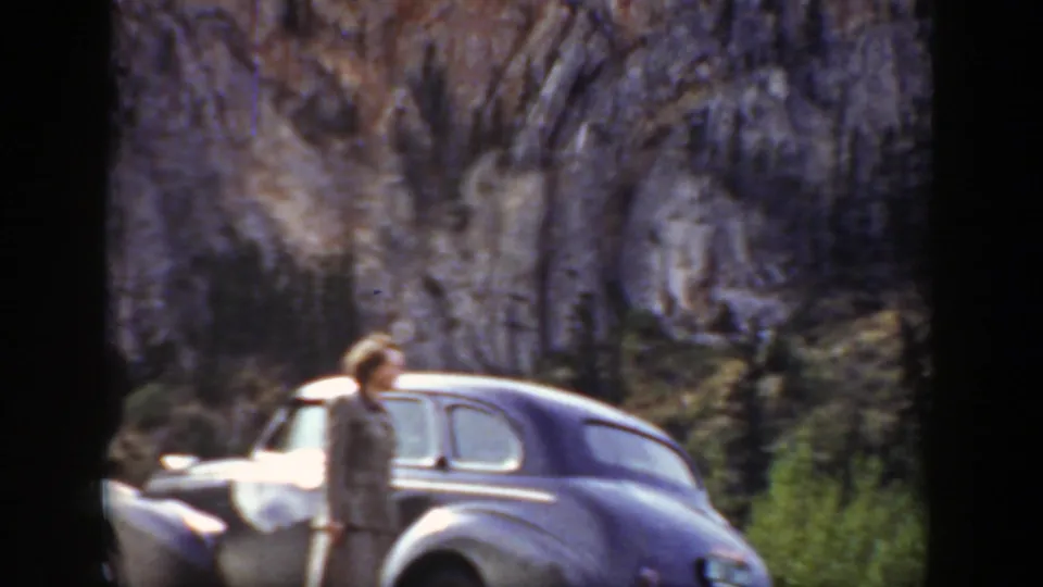 A woman stands near a parked 1930s-era sedan below a rocky Wyoming cliff in home-movie footage.