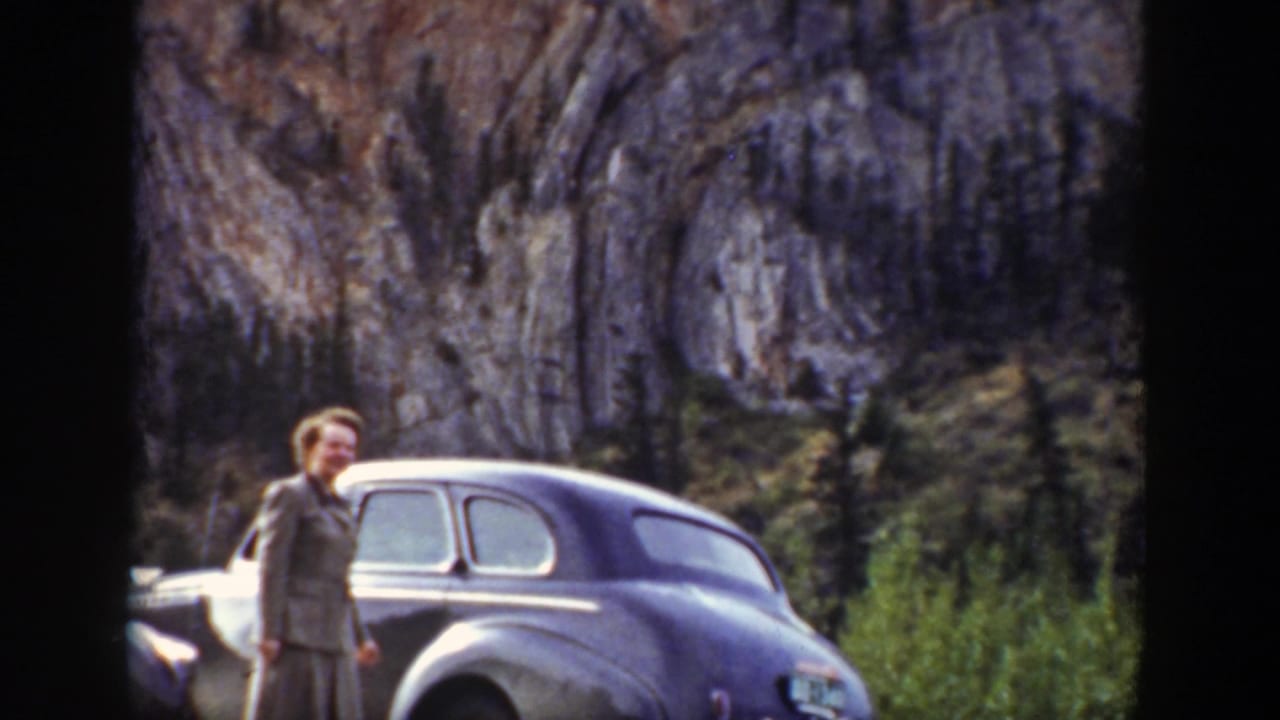 Woman standing beside a rounded sedan below rocky cliffs.