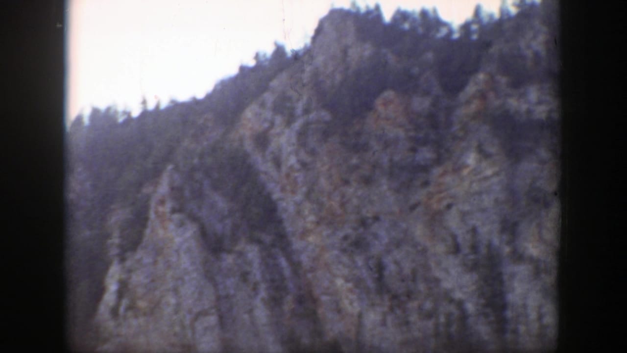 Forested rocky mountain slope under bright sky with dark film borders.