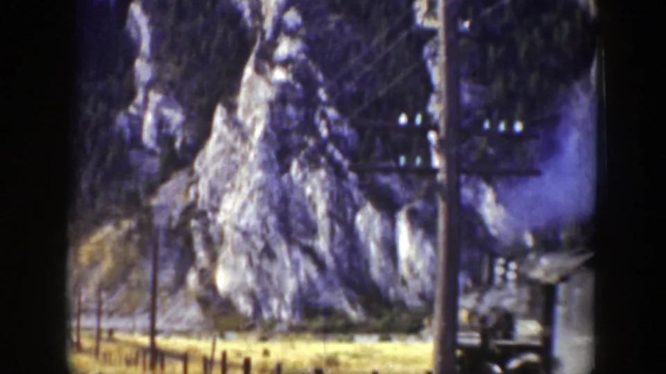 1939 Wyoming 8mm frame showing a steam train passing below rocky cliffs, with utility poles, wires, and smoke visible.